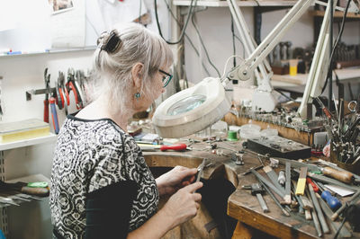 Side view of senior woman making jewelry at workbench in workshop