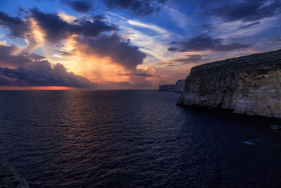 Scenic view of cliff at sea shore against cloudy sky during sunset