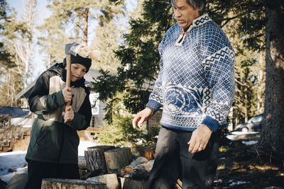 Mature man looking at son cutting log with axe in forest during winter