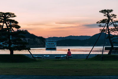 Man sitting on riverbank against sky during sunset