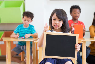 Portrait of girl showing thumbs up while sitting with slate in classroom