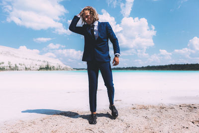 Full length of man with curly hair wearing suit while standing at beach against sky
