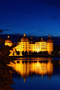 Illuminated buildings by lake against blue sky at dusk