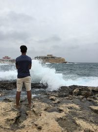 Rear view of man standing by waves splashing on rocky shore at sea against sky