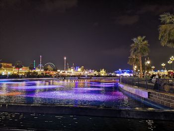 Illuminated bridge over river by buildings against sky at night