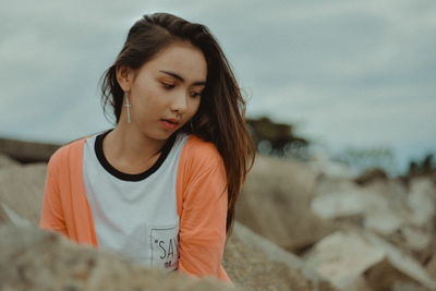 Young woman looking away while sitting by rocks against cloudy sky