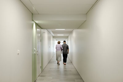 Full length rear view of senior woman walking arm in arm with male caregiver at retirement home corridor