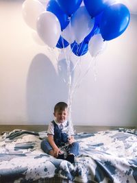 Cute boy holding balloons while sitting on bed at home