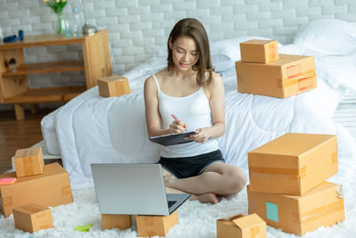 Full length of woman using laptop while sitting amidst cardboard boxes at home