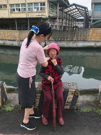 Side view of woman helping while senior person wearing hat against canal