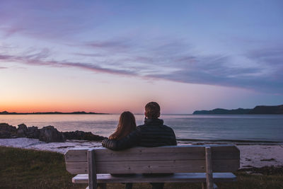 Rear view of couple sitting on shore against sky during sunset