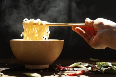 Hand picking up tasty steaming noodles with chopsticks in wood bowl and some spice on wooden table