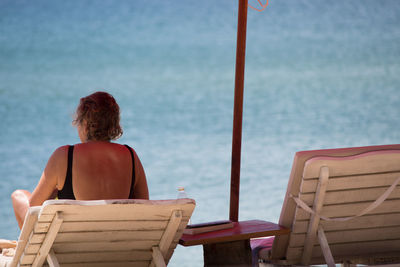 Rear view of woman in swimsuit sitting on deck chair at beach