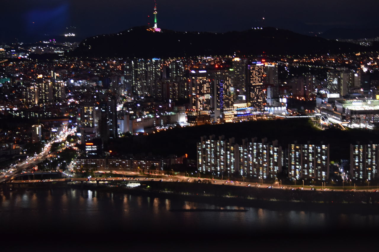 ILLUMINATED MODERN BUILDINGS BY RIVER AGAINST SKY AT NIGHT