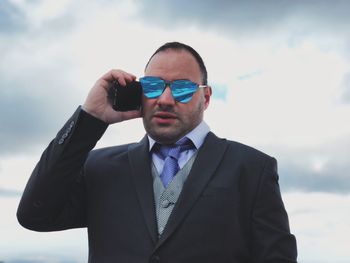 Portrait of businessman in sunglasses talking on mobile phone against sky