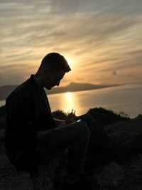 Silhouette man writing on book while sitting against lake during sunset