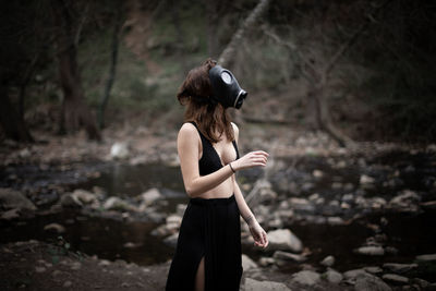 Side view of sensuous woman wearing gas mask while standing in forest