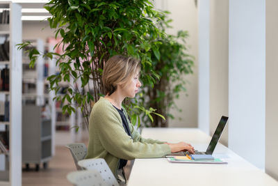 Pensive university teacher working on laptop in cozy empty library filled with green houseplants