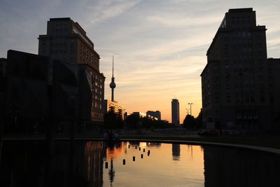 Silhouette buildings by lake against sky at sunset