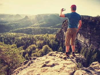 Hipster hiker. amateur takes photos with smart phone on peak of rock. dreamy spring sunrise