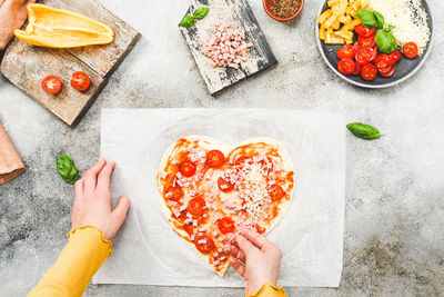 Hands of caucasian teenage girl lay down sliced cherry tomato on heart-shaped pizza dough.