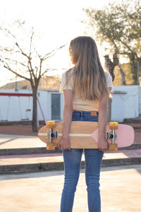 Back portrait of a slim young blonde girl holding a natural vintage skateboard behind her back