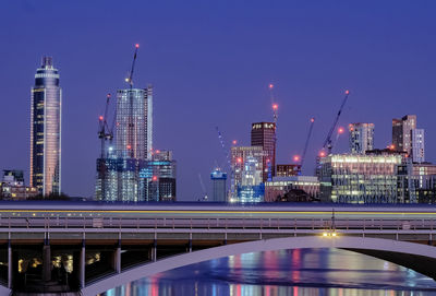 Illuminated modern buildings by river against clear sky at night
