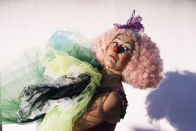 Side view of sad mature female clown in curly pink wig with bright makeup carrying pile of tutu skirts and looking away after performance on street on sunny day