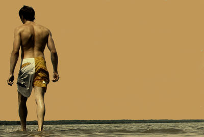 Rear view of shirtless man wading in sea against clear sky