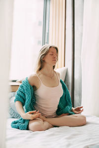 A young woman in pyjamas is sitting on a bed in a lotus position 
