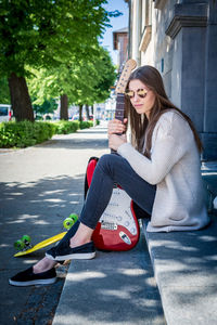 Young woman with guitar and skateboard sitting on steps in city