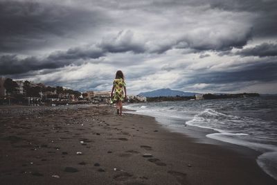 Rear view of girl walking at beach against cloudy sky