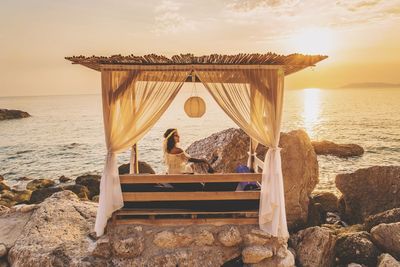 Woman sitting in gazebo on rock formation at beach against sky during sunset