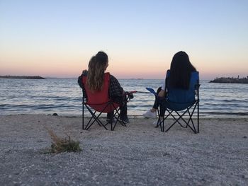 Rear view of friends sitting on chairs at beach during sunset