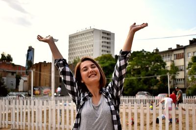 Portrait of smiling young woman with arms raised against buildings