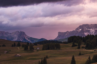Meadow in the mountains with trees and cabins against dark cloudy sky during sunset