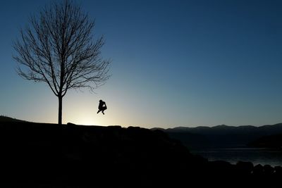 Silhouette person jumping by bare tree against clear sky