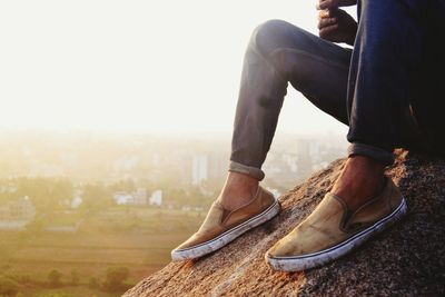 Low section of man sitting on rock against sky on sunny day