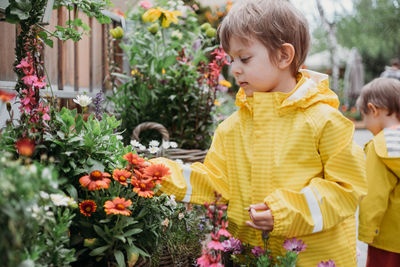 Toddler boy in yellow raincoat sniffing flowers in pots