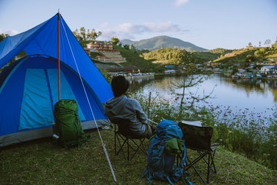 Rear view of men sitting on tent by lake against sky