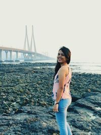 Side view portrait of young woman with bridge over sea in background against clear sky