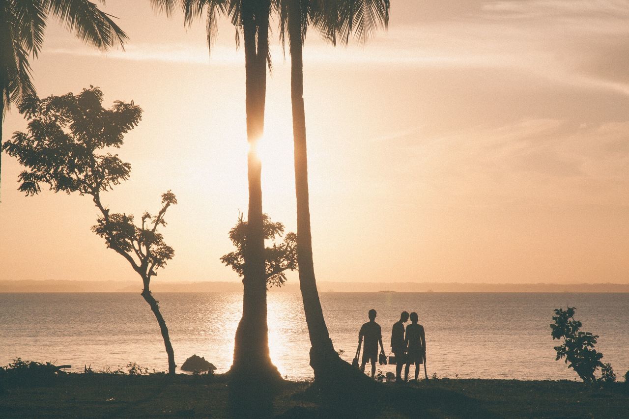 sea, horizon over water, sunset, water, silhouette, scenics, tranquil scene, tranquility, beauty in nature, shore, lifestyles, leisure activity, tree, nature, sky, idyllic, relaxation, tree trunk, orange color, sun, vacations, outdoors, tourism, unrecognizable person, ocean