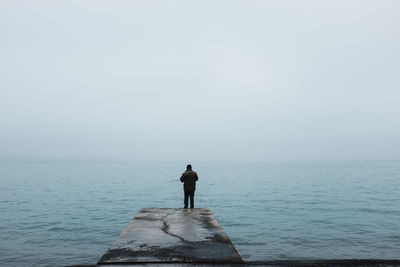 Man fishing while standing on jetty over sea against clear sky