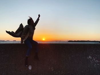 Full length of woman sitting on retaining wall by sea during sunset