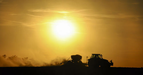 Silhouette of a tractor sowing seeds in a field in a cloud of dust against the background.