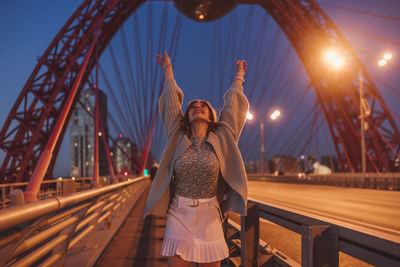 Cheerful woman standing with arms raised on bridge in city at night