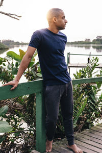 Full length of young man looking at view while standing on pier against plants and lake