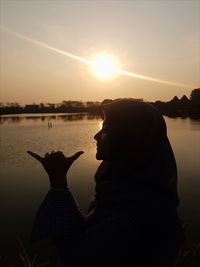 Woman gesturing shaka sign by lake against sky during sunset