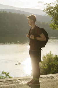 Full length of thoughtful man looking away while standing by lake at sunset