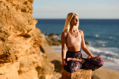 Young woman meditating while sitting on rock at beach against sky